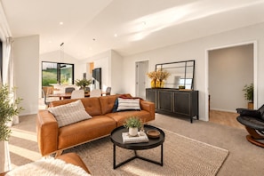 A modern living room with a leather sofa, a coffee table, and a dining area in the background. - Modern Wānaka Retreat | 3 Queen Bedrooms, Mountain Views, High Ceilings & Internal Garage (Wānaka)