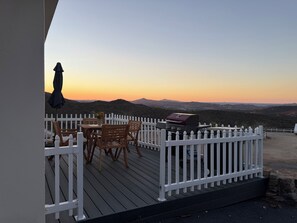 A deck with a white picket fence, wooden chairs, a table, and a barbecue grill.