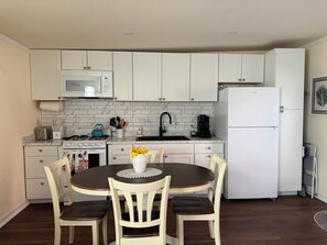 A compact kitchen with white cabinets, a microwave, a stove, a sink, and a refrigerator. There is a dining table with chairs and a vase with flowers.