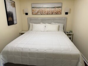 A neatly made bed with a white quilted bedspread and pillows, a headboard with a light wood finish, and a landscape painting above it.