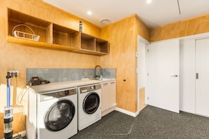 A compact laundry room with two washing machines, a sink, and a drying rack. - Modern Northlake Wanaka Home | 3 Bedrooms, Media Room, Outdoor Pizza Oven & Entertaining Area (Wānaka)