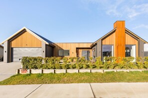 A modern two-story house with a garage, a large window, and a well-maintained garden. - Modern Northlake Wanaka Home | 3 Bedrooms, Media Room, Outdoor Pizza Oven & Entertaining Area (Wānaka)