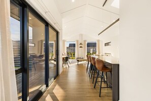 A modern interior with a bar area, seating, and a view through large glass doors. - Modern Northlake Wanaka Home | 3 Bedrooms, Media Room, Outdoor Pizza Oven & Entertaining Area (Wānaka)