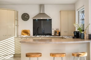 A modern kitchen with a central island, stainless steel range hood, and wooden stools. - Mountain View House | 3 Bedrooms, Sleeps 6, 2 Bathrooms, Air Conditioning & Easy Access to Lake Wānaka (Wānaka)