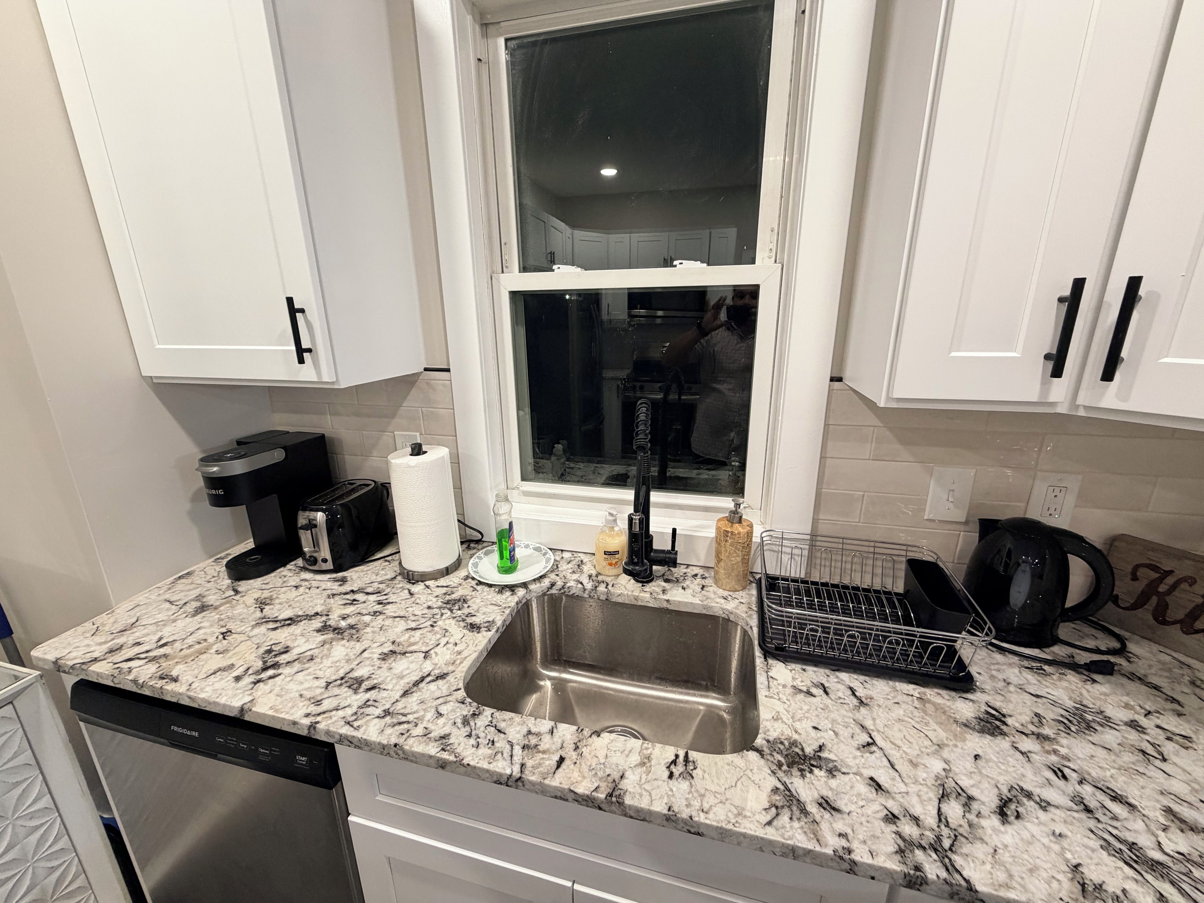 A kitchen with a stainless steel sink, a toaster, a paper towel holder, and a window with a view of a person in the background.