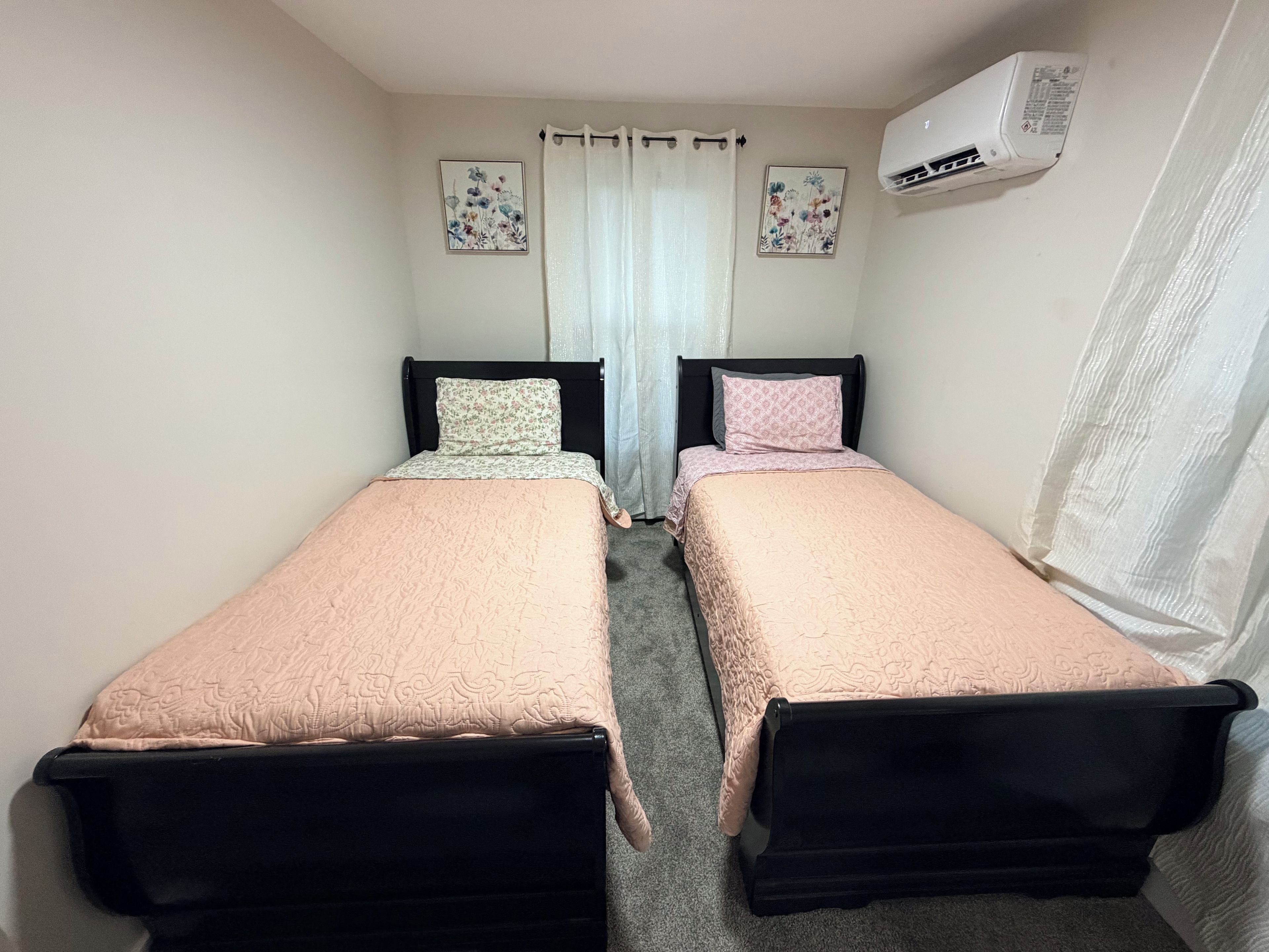 A room with two single beds, a window with curtains, an air conditioning unit, and two floral wall art pieces.