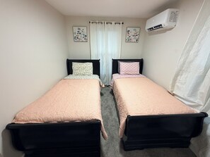 A room with two single beds, a window with curtains, an air conditioning unit, and two floral wall art pieces. - Delightful 4-bedroom house in Carneys Point Township, near Wilmington & Philly! (Carneys Point Township)