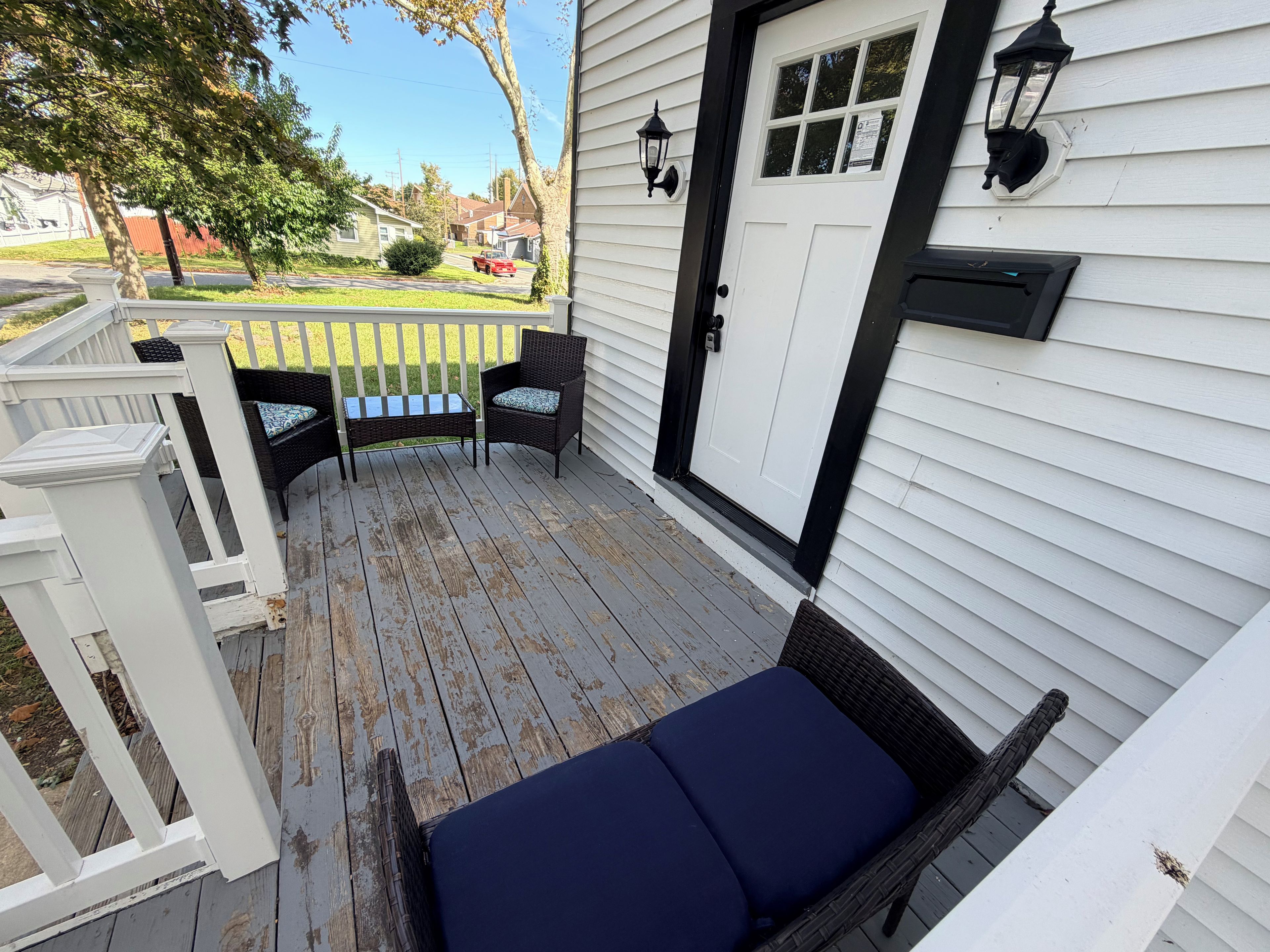 A covered porch with a bench and chairs, a white door, and a view of the neighborhood.