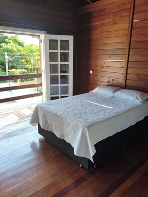 A wooden room with a bed, a sliding glass door leading to an outdoor area, and a view of greenery outside. - House with Pool in Bombinhas (Bombinhas)