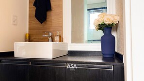 Ein Badezimmer mit weißem Waschbecken, einem Spiegel, einer blauen Vase mit Blumen und einem dunklen Holzschrank mit einem Logo.