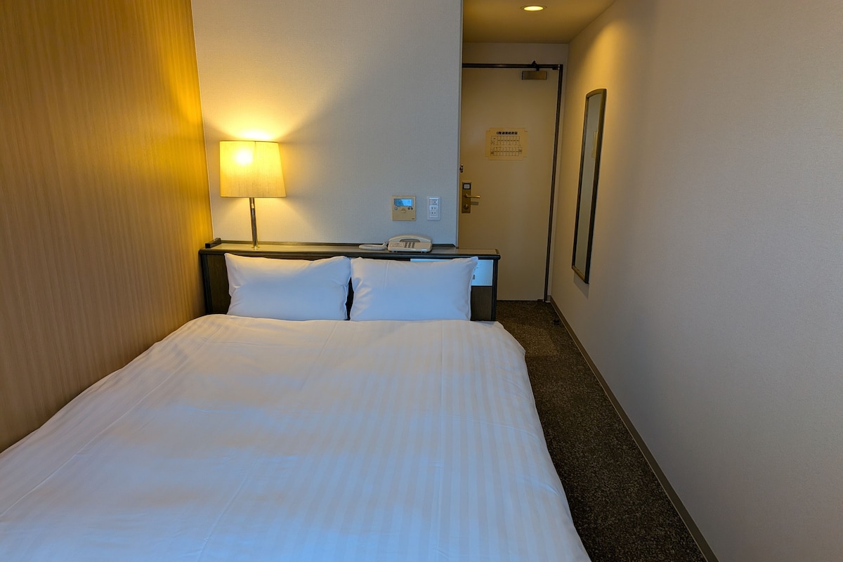 A hotel room with a bed, a bedside lamp, a phone, and a door leading to another room.