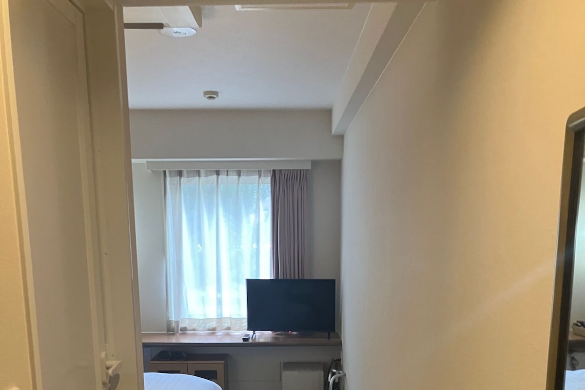 A hotel room with a bed, a desk with a television, a window with curtains, and a carpeted floor.