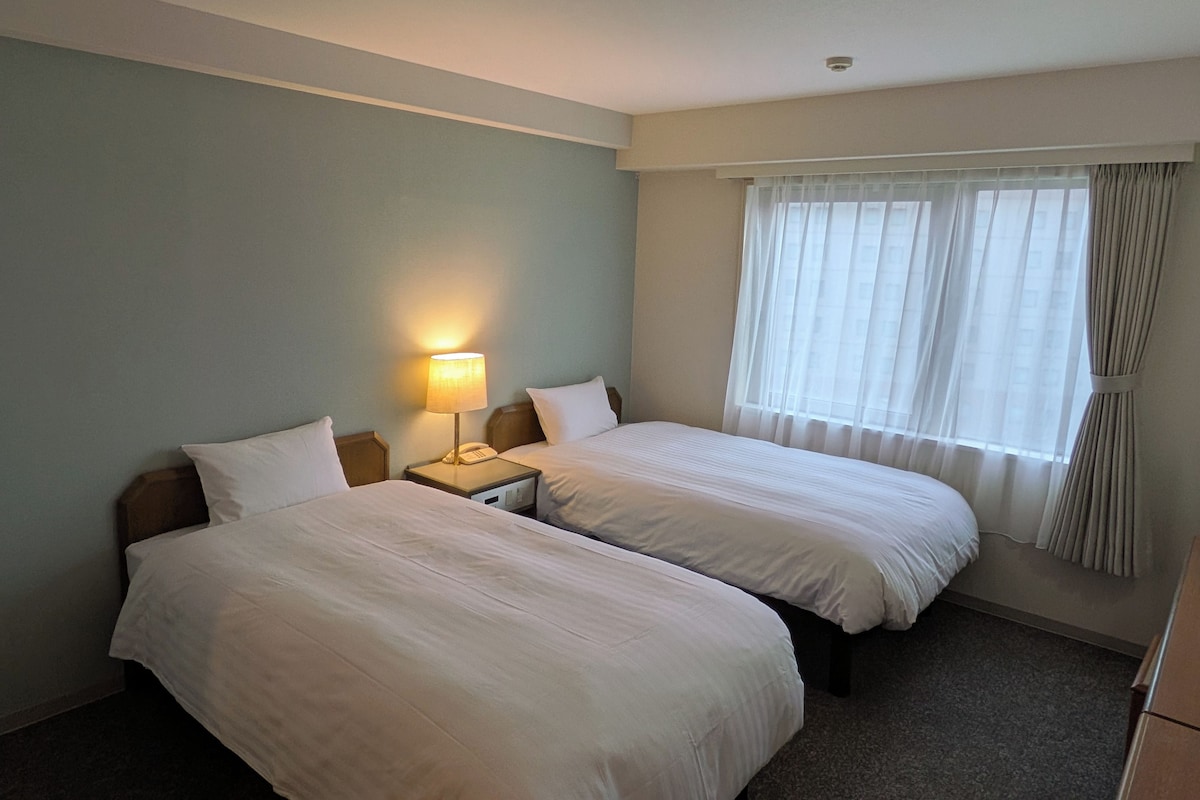 A hotel room with two beds, a nightstand with a lamp, and a window with curtains.