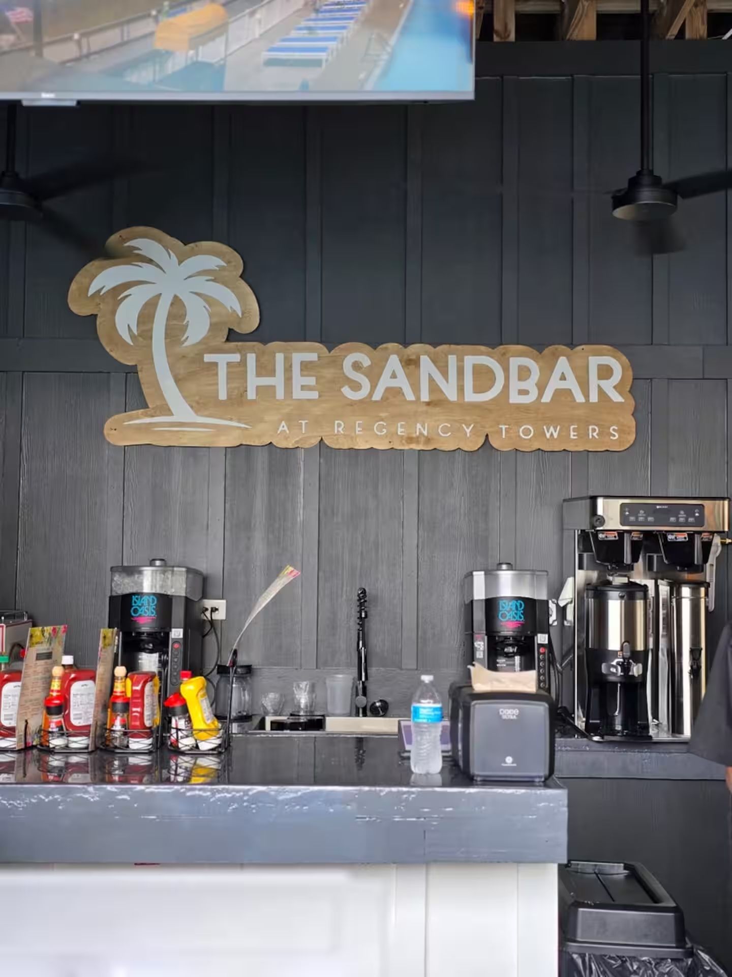 A bar area with a sign reading 'The Sandbar at Regency Towers,' coffee machines, condiments, and a water bottle on the counter.