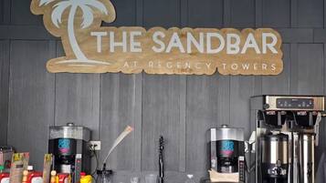 A bar area with a sign reading 'The Sandbar at Regency Towers,' coffee machines, condiments, and a water bottle on the counter.
