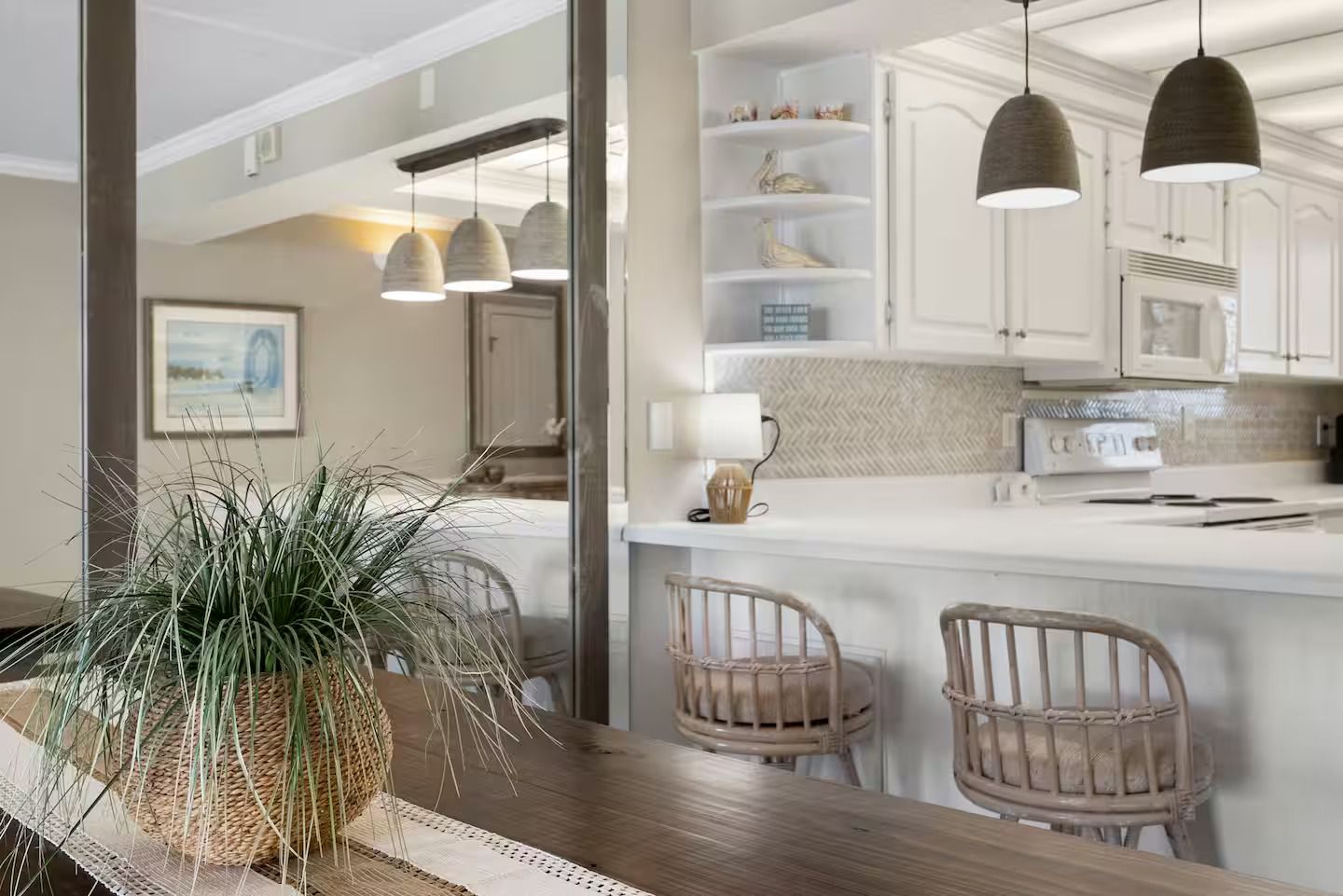 A kitchen with white cabinets, a white countertop, and pendant lights. There is a plant on the table and wicker chairs.
