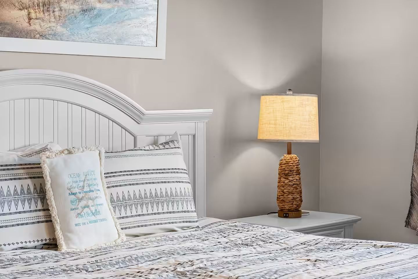 A neatly made bed with patterned bedding, a bedside lamp with a woven base, and a wall-mounted painting.