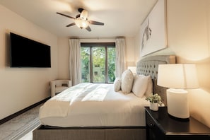 Ein Hotelzimmer mit einem großen Bett, einem Flachbildfernseher, einem Deckenventilator, einem Fenster mit Vorhängen und einer Nachttischlampe.