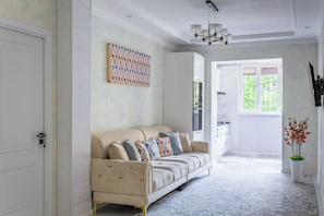 A modern living room with a beige sofa, patterned cushions, a chandelier, and a large window with a view of greenery. - Apartments in the historic center (Tashkent)