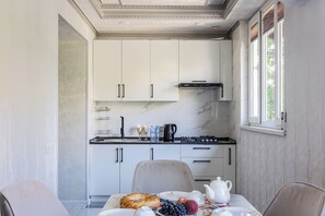 A modern kitchen with white cabinetry, a black stove, and a window with blinds. - Apartments in the historic center (Tashkent)