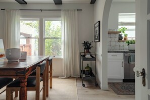 A dining area with a wooden table and chairs, a kitchen with white cabinets, and a view of greenery outside. - Casa Mika - Designer Curated 2BR/1BA Retreat Near SoFi, Intuit Dome & The Forum (Los Angeles)
