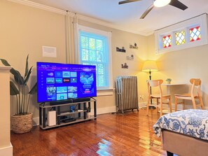 A living room with a television displaying a Roku TV interface, a radiator, a ceiling fan, a potted plant, and a dining area with a table and chairs.