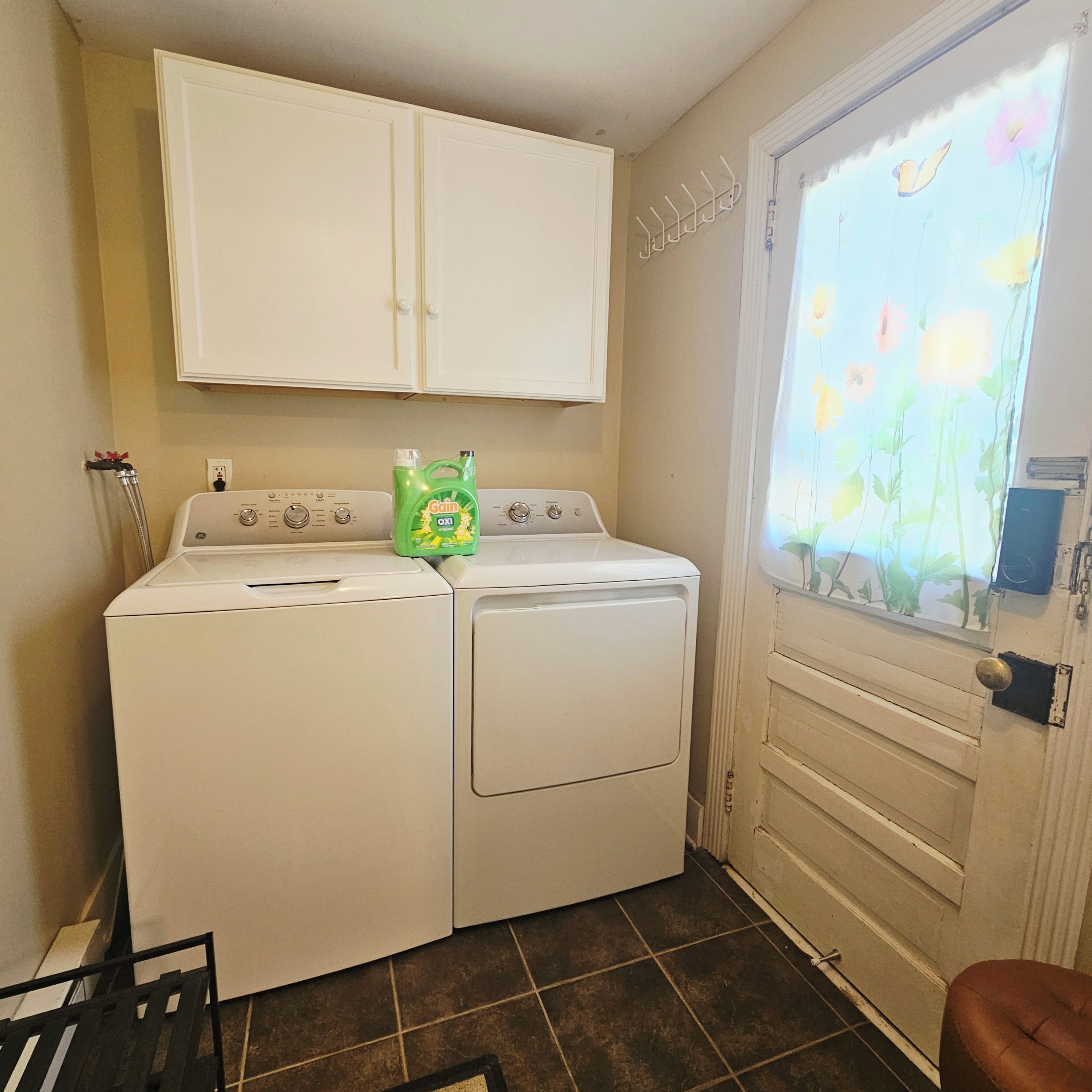 A laundry room with a front-loading washer and dryer, a window with a floral view, and a white cabinet above the appliances.