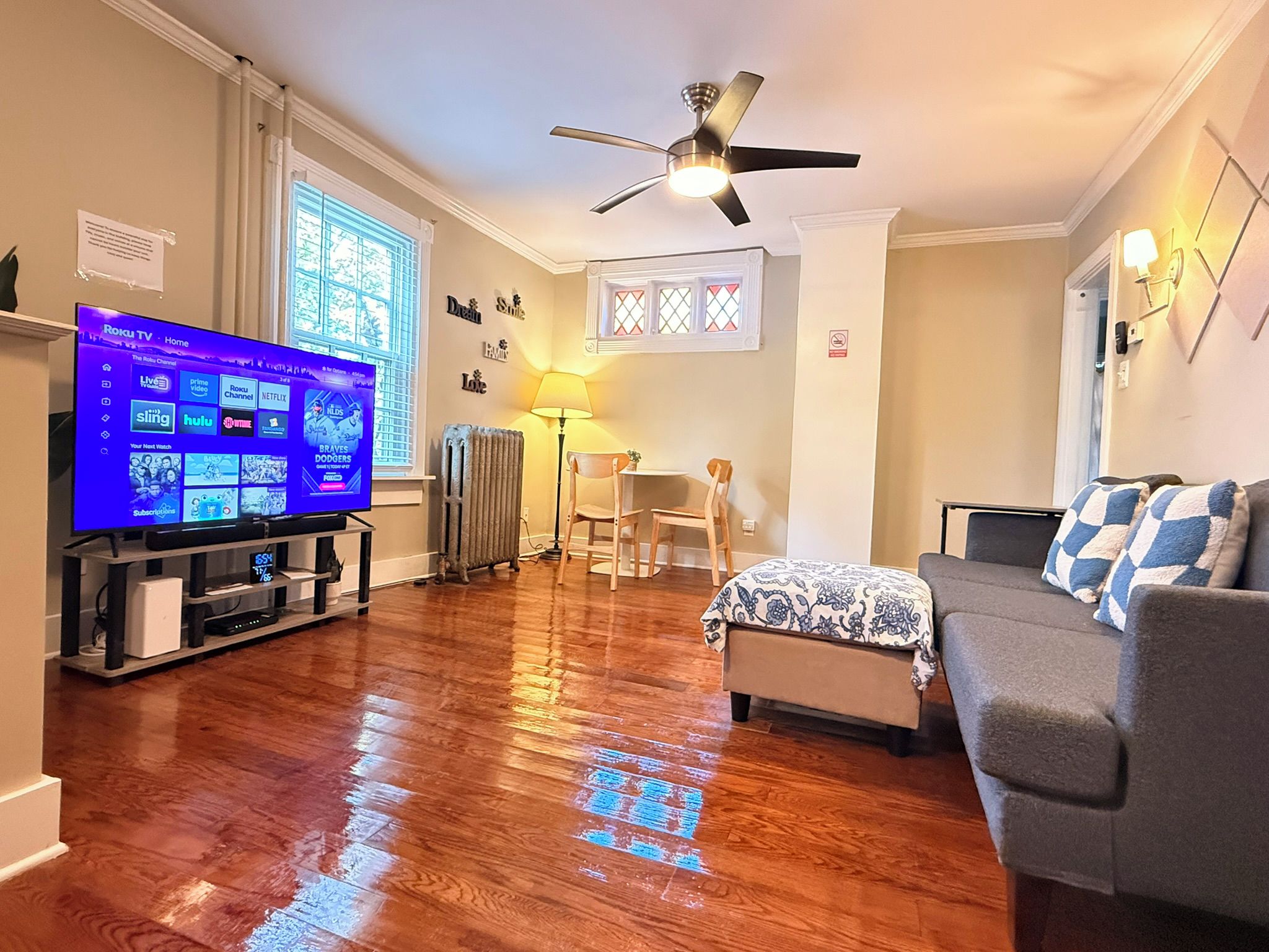 A living room with a flat-screen TV, wooden flooring, a ceiling fan, a sofa, and a window with blinds.