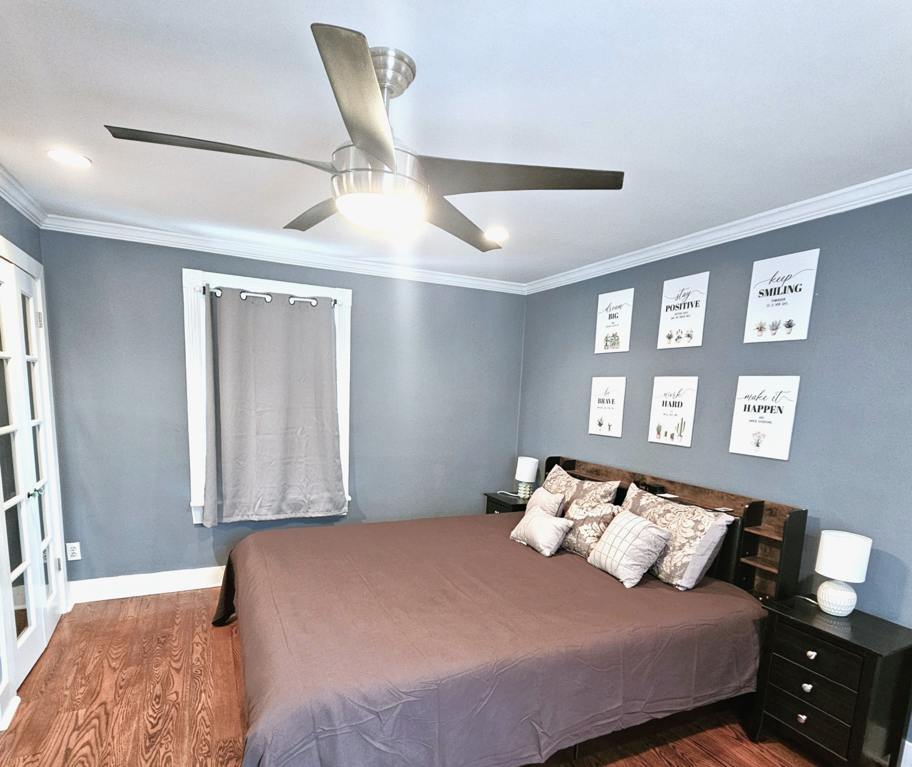 A bedroom with a bed, a ceiling fan, a window with a curtain, and motivational wall art.