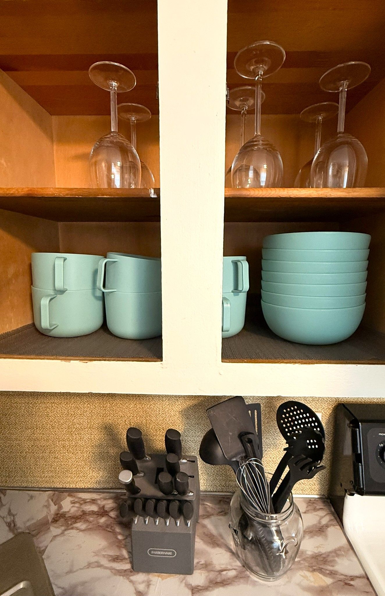 A kitchen shelf with wine glasses, cups, and a spice rack.