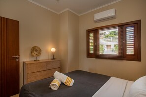 Een hotelkamer met een bed, nachtkastje met lamp, houten hoofdbord, raam met zonwering, en een airconditioning.