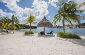 A beach with palm trees, a thatched umbrella, and lounge chairs. - La Marina | 2BR Waterfront + Marina Views (Brakkeput)