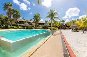 A swimming pool with a sandy beach area, palm trees, and thatched umbrellas. - La Marina | 2BR Waterfront + Marina Views (Brakkeput)