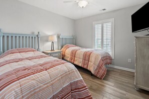 A bedroom with two beds, a nightstand, a lamp, a radiator, a television, and a window with blinds. - Sea Oats | 3BR Oceanfront Escape (North Myrtle Beach)