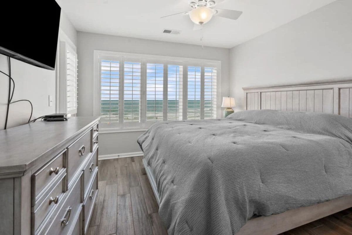 A bedroom with a large bed, a dresser, a ceiling fan, and a window with a view.