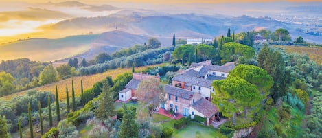 A villa with multiple buildings, surrounded by lush greenery and vineyards, overlooking rolling hills and a valley.