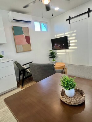 Habitació moderna amb taula de fusta, un sofà gris, televisió penjada a la paret i una planta en test.