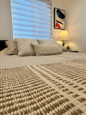 A bed with a textured blanket, a wall-mounted lamp, and a window with blinds. - LUUM House (Phoenix)