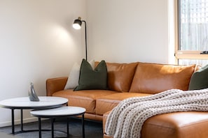 A room with a brown leather sofa, two small round tables, a floor lamp, and a window with blinds. - The Mersey Luxe Retreat (East Devonport)