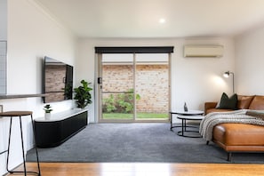 A modern living room with a sofa, a TV, a coffee table, and a sliding glass door leading to a brick exterior. - The Mersey Luxe Retreat (East Devonport)
