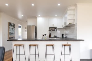 A modern kitchen with white cabinets, a black countertop, and four wooden stools. - The Mersey Luxe Retreat (East Devonport)