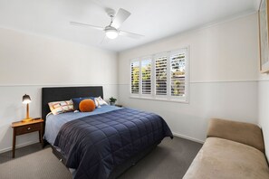 A bedroom with a bed, nightstand, lamp, ceiling fan, window with blinds, and a chair. - Renovated on Elizabeth (Anglesea)