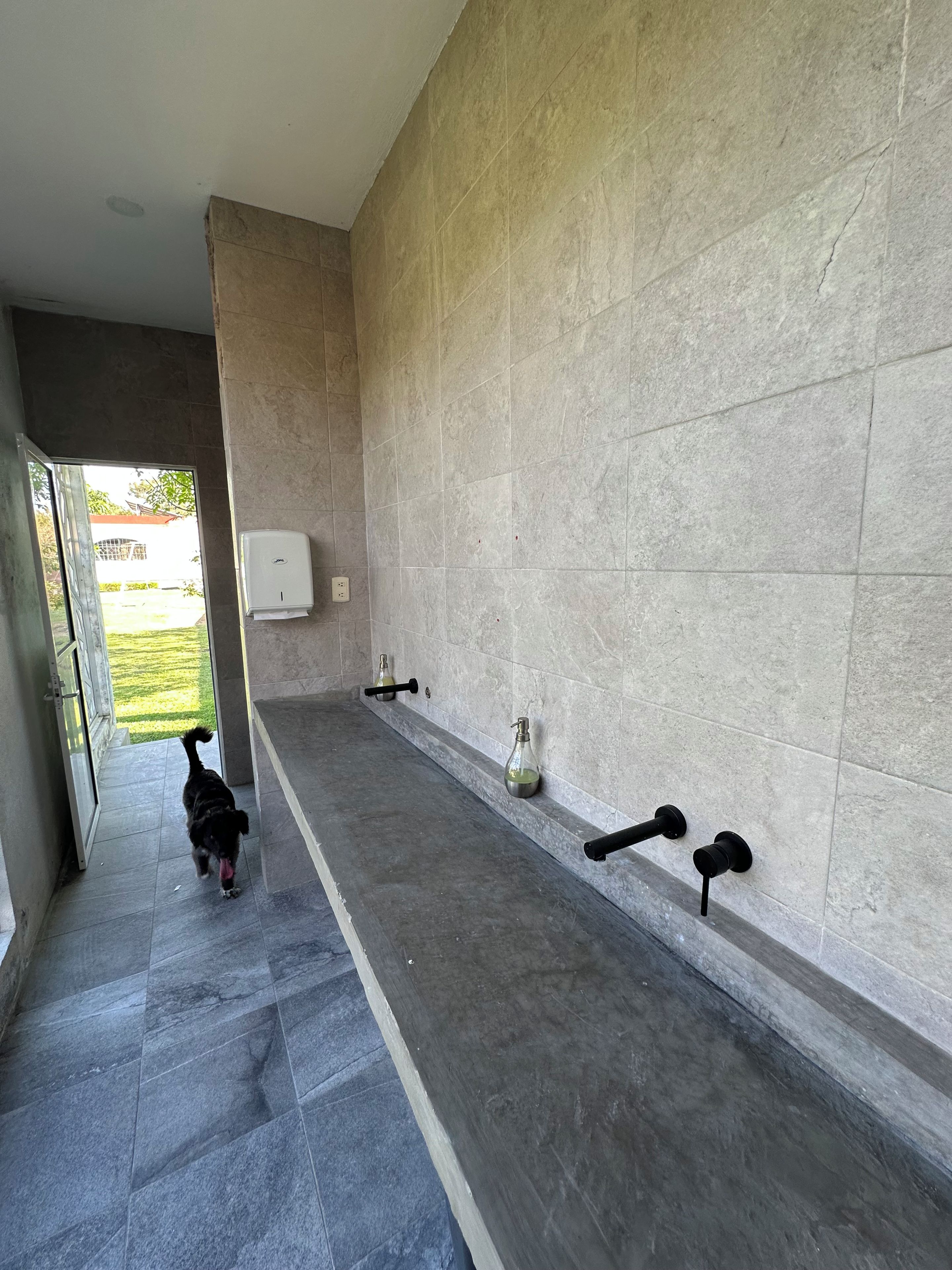 Un pasillo con un perro caminando hacia una puerta, una barandilla y un secador de manos fijado a la pared.