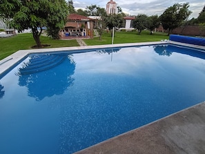 A swimming pool with a clear blue surface, surrounded by a grassy area and a building with a red roof. - Country Cottage in Morelos · Private Pool · Pet-Friendly (Fraccionamiento Paraíso Tlahuica)