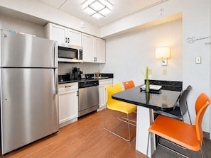 A compact kitchen with stainless steel appliances, white cabinets, and a dining area with orange chairs. - Dallas Stay | Pool + Gym | Near Love Field & UTSW (Dallas)