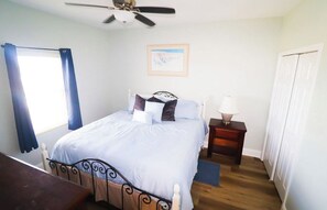 Ein Schlafzimmer mit einem Bett, einem Nachttisch, einem Deckenventilator, einem Fenster mit Vorhängen und einer Tür.