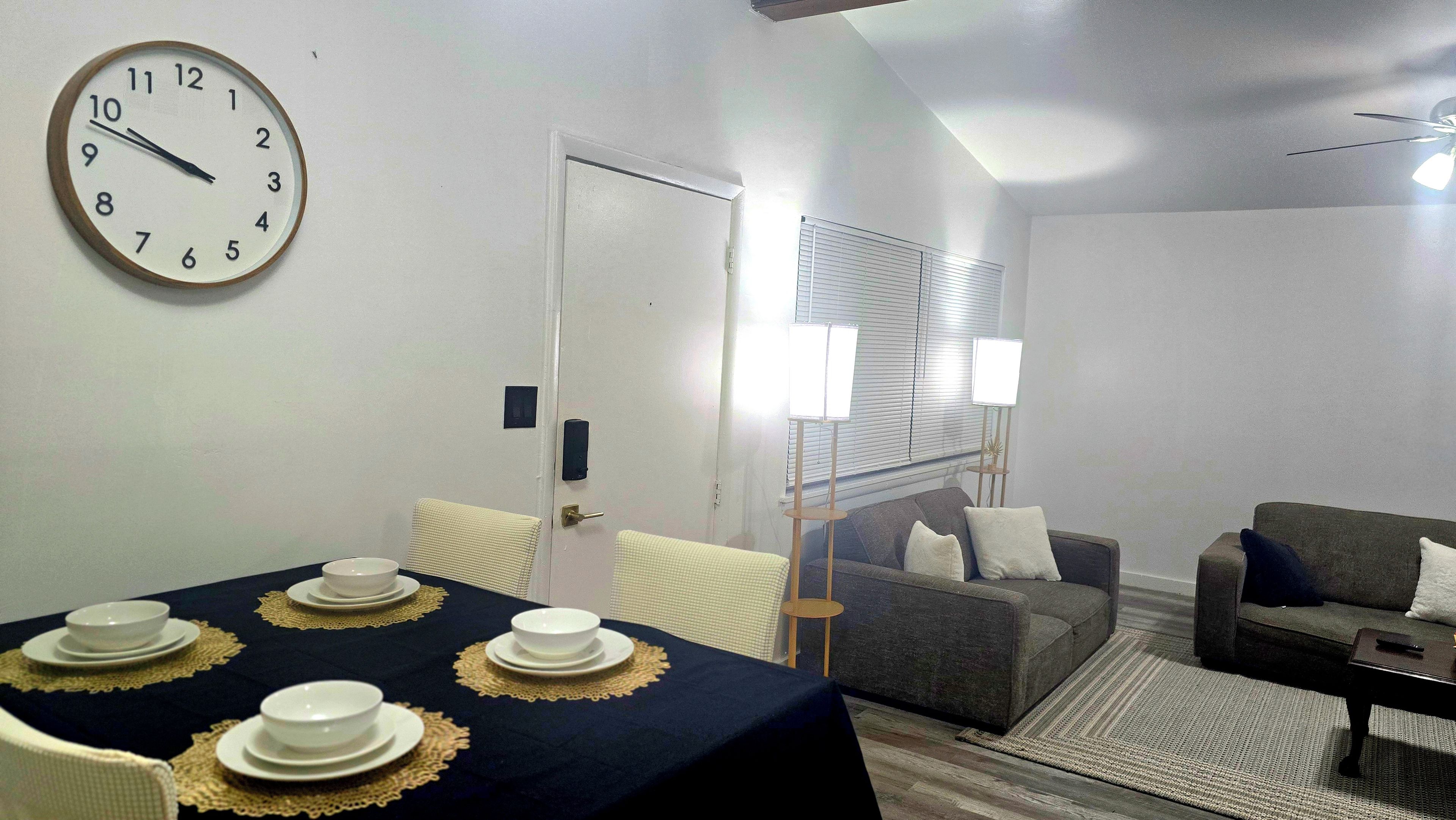 A room with a dining table set for four, a sofa, a chair, and a clock on the wall.