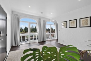 A spacious room with large windows, a view of greenery, and a white textured wall with framed artwork. - The White Terraces - Spa Pool, Sleeps 10 (Rotorua)
