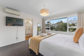 A hotel room with a bed, a television, a window with curtains, and a view of greenery outside. - Huia Nest - Walk to Golf & Stadium, Sleeps 8 (Rotorua)