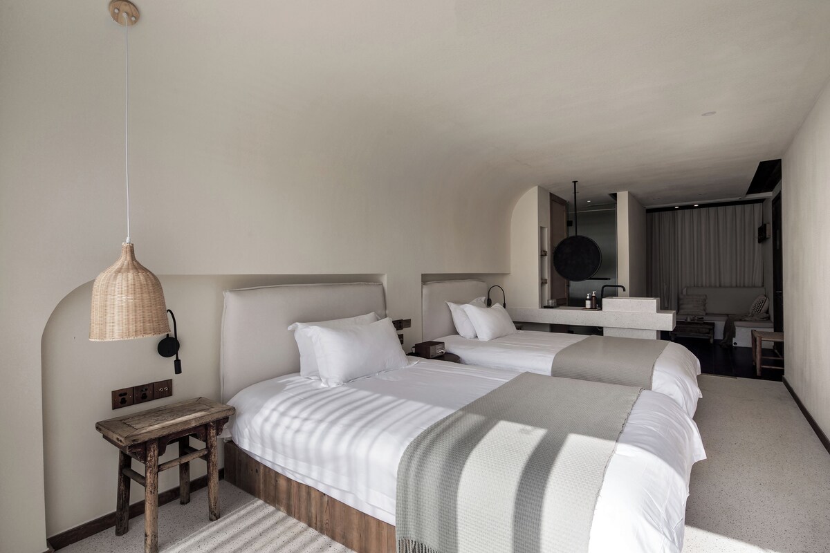 A hotel room with two beds, a wooden nightstand, a woven lamp, and a bathroom with a mirror and sink.
